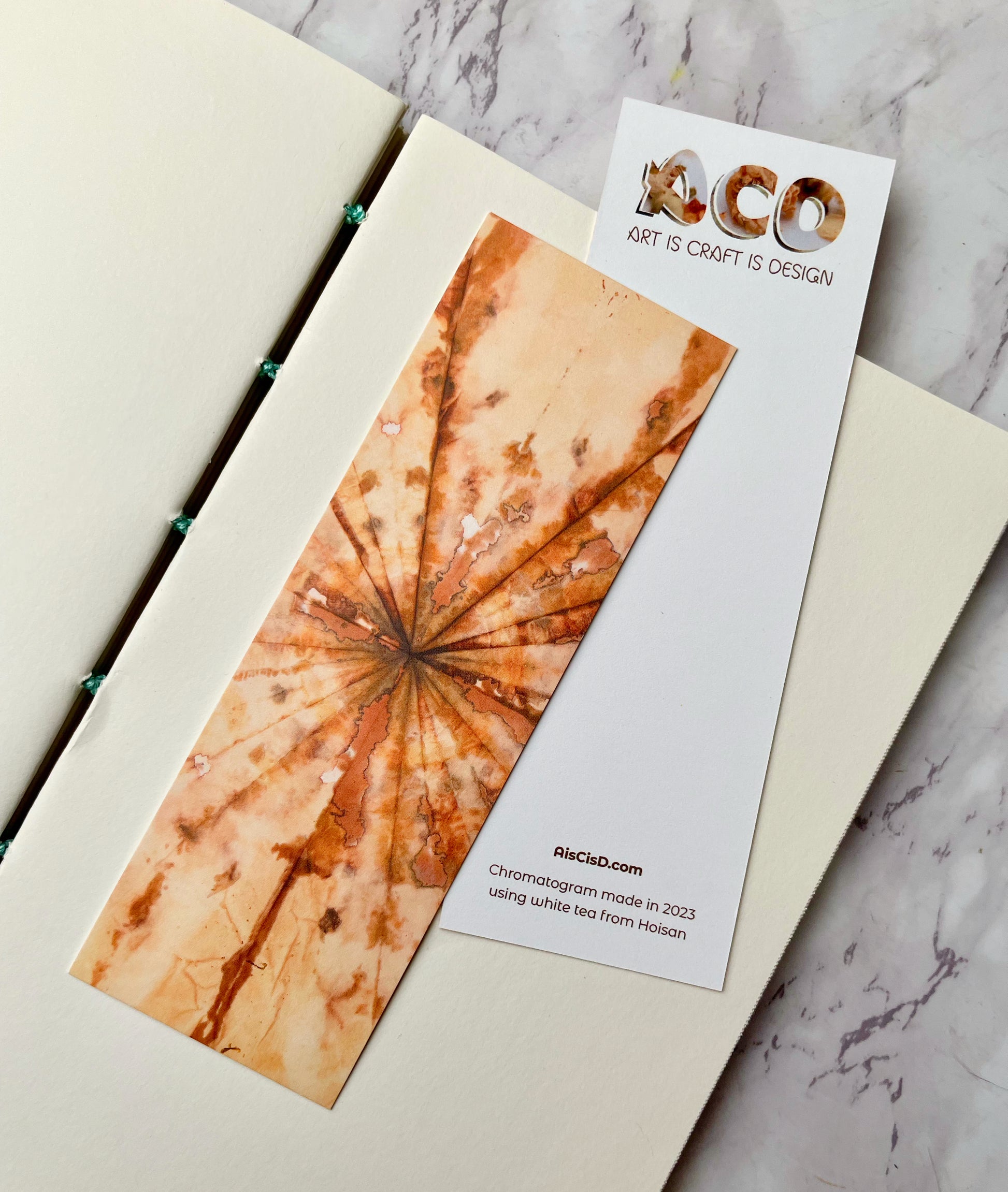 A bookmark on top of an open book. It has tans and browns radiating out from a central point. It's on top of another bookmark that shows the Art Is Craft Is Design logo and has text that says: AIsCIsC.com Chromatogram made in 2023 using white tea from Hoisan.