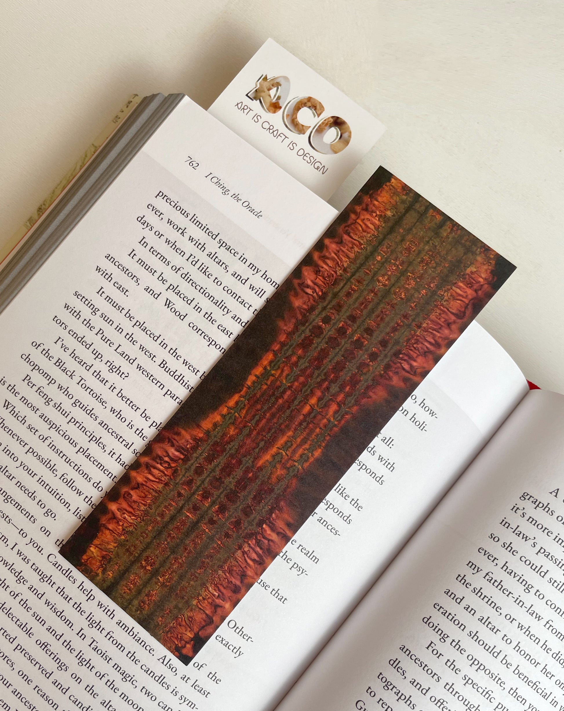 A bookmark on top of an open book. It shows bursts of red radiating out from five dark lines running across the length of the paper. The top of another bookmark is sticking out of the book and shows the Art Is Craft Is Design logo.