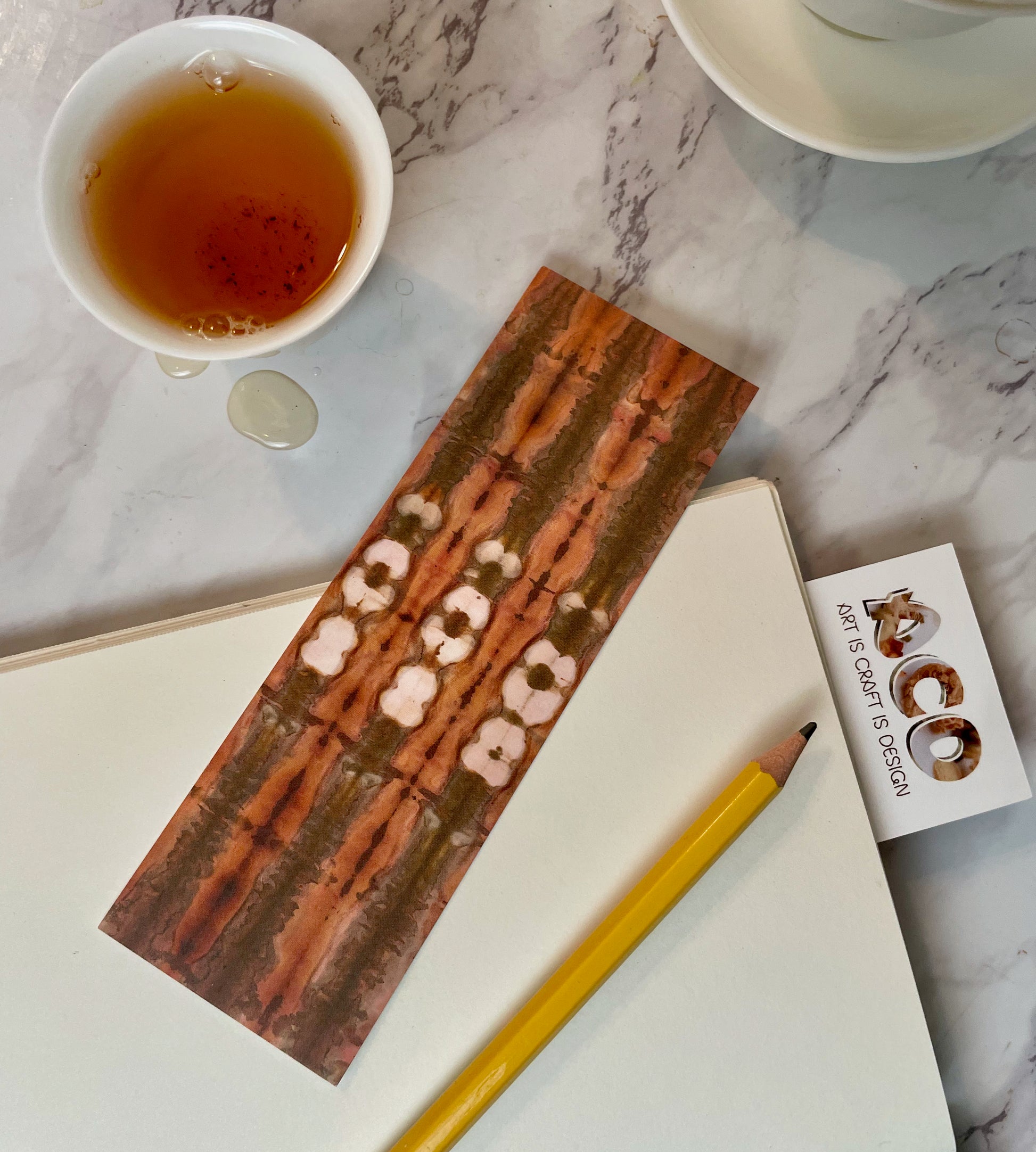 A bookmark on top of an open journal with a pencil and next to a cup of tea and a gaiwan. The bookmark has brown and light red uneven and wide lines running along its length with three sets of white splotches interrupting the darker lines. Sticking out of the journal is the top of a bookmark showing the Art is Craft is Design logo.